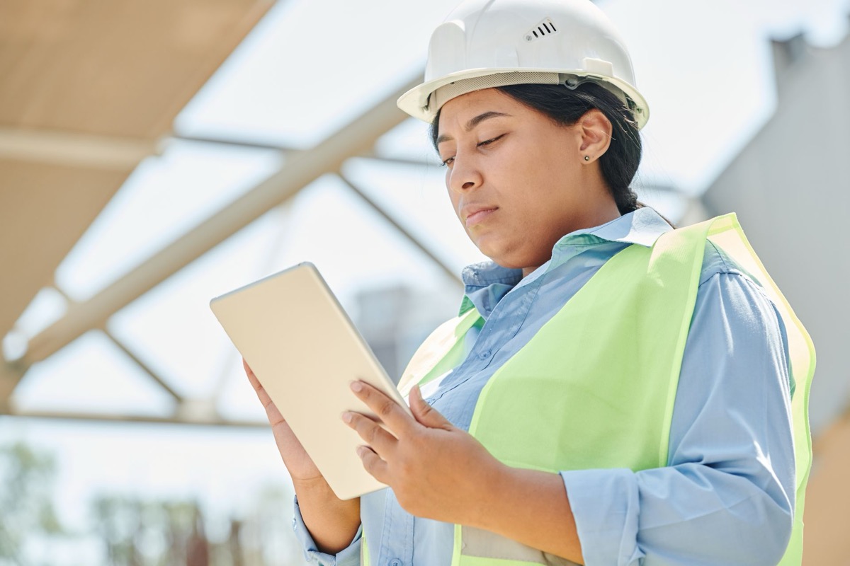A construction worker in a hard hat and hi-vis vest reading on a tablet